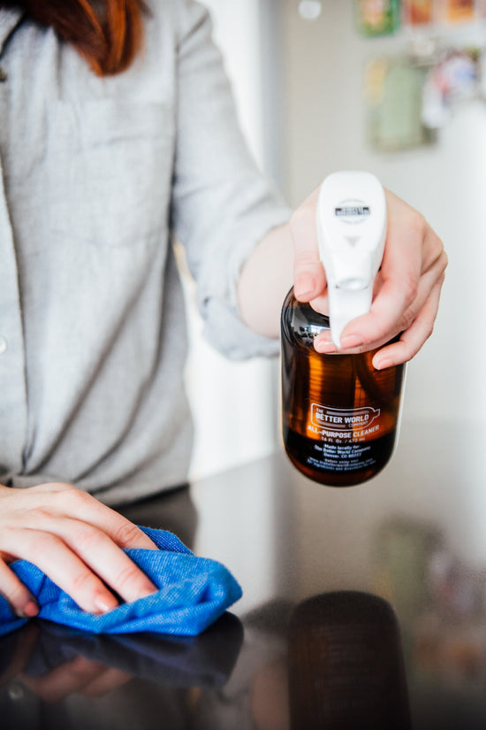 All-Purpose Cleaner sprayed from a reusable amber glass bottle onto a countertop and wiped with a microfiber cloth.