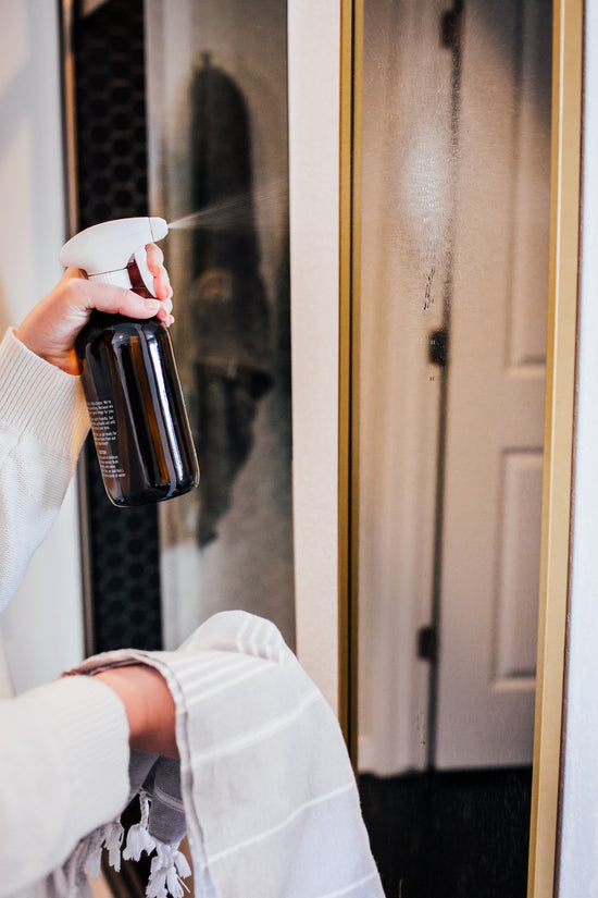 Glass Cleaner sprayed from a reusable amber glass bottle onto a glass mirror while wiping with a cloth.